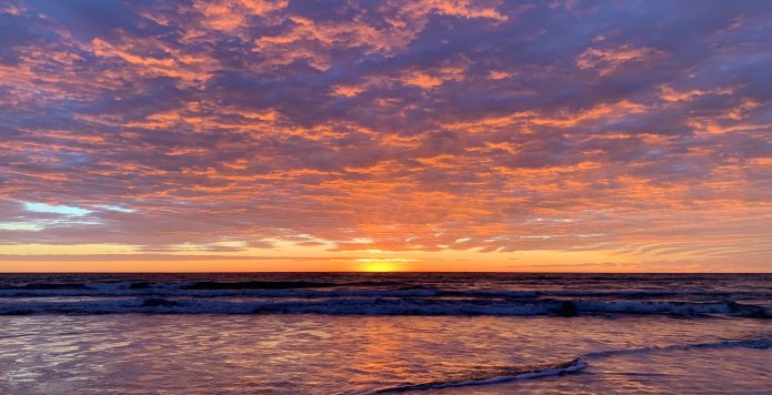 Sunrise Over The Sea From Manly Beach. The Sky Is Dotted With Clouds That Seem To Glow Orange, Pink And Mauve.